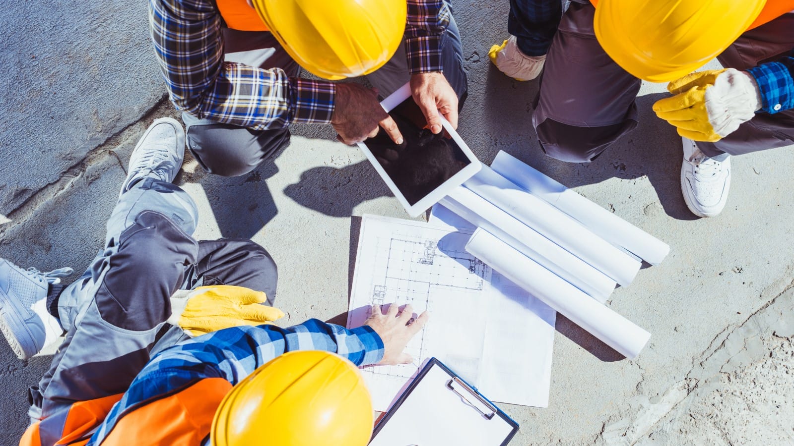 Top-down view of engineers in hard hats reviewing blueprints and a digital tablet on a construction site.